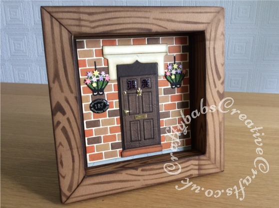 New Home Shadow Box card made using a variety of dies including Marianne bricks die (comes with embossing folder), XCut georgian door die set (cut into for over door stone and step), Spellbinders nesting ovals die for name plaque, bird cage die snipped for hanging baskets. Nesting rectangles die for door scored on score board to create moulded door panels, Tattered lace small flowers die. Glossy accent used on windows in door and house name plaque. Frame made using template created from a box that Christmas cards came in, inked with distress inks through a wood grain stencil. Frame stand cut using Sizzix Bigz Tim Holtz small easel die - craftybabscreativecrafts.co.uk