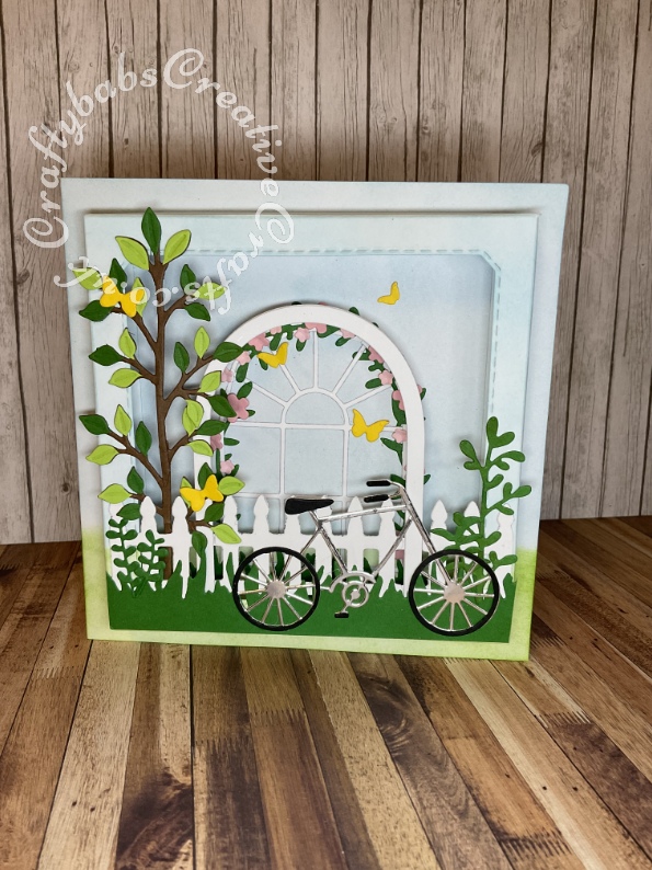 3D shadow box effect card made using various dies including: Surprise Creations Double stitched nested square dies for aperture, Cheery Lynn Designs FRM142 Window B and Sizzix Framelits Nested Arch dies for flower, arch, Go Kreate Fence die, Xcut English Countryside bprders grass border die, Creative Expressions Sue Wilson Finishing Touches Spring Foliage Dies, Memory box butterflies die, Memory box Tandem die, cut down to make bicycle, and various small foliage dies from Sissix thinlits gardening themed die sets. - craftybabscreativecrafts.co.uk